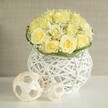 A contemporary yet romantic wedding centerpiece is shown as if arranged for a stylish reception in Queensbury, perhaps in a boutique venue not far from Queensbury Underground Station. At its heart is a compact, dome-shaped bouquet of pale butter-yellow roses, so closely clustered that their velvety petals form a seamless mound of colour. Each rose is fully open, revealing layered spirals that hint at a gentle, sweet fragrance. Between the roses, airy sprays of white baby's breath thread through the design, their tiny round blossoms creating a soft halo effect, like a fine mist drifting over a garden at dawn. The interplay of creamy yellow and crisp white produces a light, uplifting palette that feels both elegant and approachable.

A narrow collar of rich green foliage hugs the base of the bouquet, its sleek leaves arranged with precision to create a defined edge where the flowers meet the container. This container is a sculptural white woven vase, rounded and slightly cinched at the waist, made from slender strands that interlace to form an open, basket-like lattice. The structure reads as modern and airy, allowing glimpses of the stems within and casting delicate, crisscrossing shadows onto the smooth white table surface below. Placed beside the vase are two decorative spheres crafted from pale, rattan-style strips, their open geometric construction echoing the vase's woven texture and giving the scene a harmonious, curated look.

Behind the arrangement, a softly blurred wall in warm beige tones provides a calm, neutral backdrop, similar to the soothing interiors of many Queensbury flats and family homes. The image conveys a sense of gentle celebration and polished minimalism, ideal for wedding tables, anniversary lunches, or intimate gatherings where Queensbury hosts want flowers that feel fresh, local, and quietly refined.

