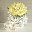 Creamy yellow roses are gathered into a compact dome, their ruffled centers opening like gentle sighs beneath a halo of tiny white gypsophila that veils the composition in a soft, cloudlike breath. Each rose retains a poised, upright form-petals layered with a velvet softness that catches the morning light-while the baby's breath whispers between blooms, lending a feathery rhythm and a hint of translucence. The vessel is a striking white wicker orb with an airy, architectural presence: its woven ribs create shadows and depth that echo the bouquet's balance of structure and softness. A pair of minimalist woven spheres sits nearby as deliberate, modern accents, their clean geometry playfully contrasting the organic curves of the flowers. The olfactory impression is of fresh-cut fields on an early-summer day, where a faint earthiness underpins the sweetness of petals and gypsophila; it is clean, linen-like, and quietly nostalgic. Small curled blades of green arc around the edge, providing a subtle graphic frame that keeps the gaze contained and calm. This composition, described in the voice of a Queensbury florist, feels ideal for a gentle congratulation, a serene gathering, or as a wedding accent balanced between contemporary design and classic floral language. Available for local delivery across Queensbury and neighbouring Kingsbury, it carries the kind of calm refinement that makes a room feel composed and cared for.