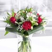 A striking red and white bouquet stands proudly in a clear cylindrical glass vase, its stems neatly trimmed and visible through the fresh water, like a window into the florist's careful craftsmanship. Deep red roses with velvety petals begin to unfurl at the heart of the arrangement, paired with a large, ruffled crimson carnation that draws the eye with its intricate layers and soft, almost feathery texture. Around them, smaller red blooms echo the same passionate tone, creating a rich, romantic core that feels perfect for an anniversary surprise or a special celebration in Queensbury. Bright white alstroemeria, with slim petals and fine speckles, weave between the reds, joined by creamy white blossoms and several closed white lily buds whose pale green tips hint at flowers soon to open. Wisps of delicate gypsophila float through the design like tiny stars, giving the bouquet an airy lightness and gentle movement. The blooms are framed by lush greenery in multiple tones: broad, glossy dark green leaves are curved and wrapped at the base to form an elegant collar, while more slender, eucalyptus-like stems add height, texture, and a touch of woodland freshness. The whole piece rests on a pristine white surface, set against a softly blurred, bright background that suggests daylight streaming through a window on a quiet Queensbury street. The lighting is even and soft, catching the moisture on the foliage and the subtle sheen of each petal, creating a mood of warmth, sincerity, and thoughtful care-an arrangement that feels at home on a dining table, office desk, or front room sideboard anywhere in the local area.