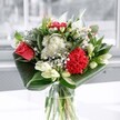 A contemporary glass-vase arrangement photographed in soft daylight, featuring a single coral-red rose and a plush crimson carnation surrounded by clouds of delicate white blooms, as crafted by a local Queensbury florist. The rose, positioned slightly off-centre, opens generously, its layered petals almost velvety, while the ruffled carnation beside it adds depth and texture with its rich, passionate tone. Encircling these focal flowers are clusters of small white chrysanthemums and daisy-like blossoms, their rounded, cushiony heads creating a gentle, almost misty halo of petals. Slender stems of baby's breath weave between them, introducing tiny star-shaped flowers that give the bouquet a light, airy quality. Creamy white lilies, mostly in bud with just the tips of their petals beginning to part, rise gracefully above the lower flowers, suggesting new beginnings and quiet anticipation. At the base, deep emerald leaves are folded and wrapped around the stems in wide, glossy arcs, lending a modern, sculptural edge that frames the arrangement and emphasises its silhouette. The clear glass vase reveals the neat, spiral stems and clean water, catching glints of light from a nearby window, much like the calm brightness streaming into a home off Queensbury Circle. The overall mood is uplifting yet composed, ideal for marking an important milestone, welcoming a new baby, or simply bringing a bright, fresh presence to a living room or office in and around Queensbury, where the crisp fragrance of lilies and greenery evokes a cool morning stroll through the local parks.