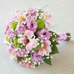 Presented against a soft, neutral-toned fabric, this vibrant and rounded bouquet overflows with a harmonious blend of lilac, blush pink, chartreuse green, and pure white tones-celebrating both contemporary taste and classic elegance, as found in Queensbury's beloved gatherings. At the heart are full, silky roses in graduated lavender and pink, unfurling with grace and inviting a gentle touch. Intertwined throughout are clusters of lime-green button chrysanthemums, adding modern flair and vivid contrast evocative of freshly tended gardens near Queensbury Station. Pink alstroemeria lilies contribute an artistic effect, their tapered petals delicately marked with vivid splashes of yellow and crimson. Dainty sprigs of baby's breath weave between the larger blooms, softening the overall vision and casting a light, ethereal veil, suggestive of celebration and hope. Beneath, dark green leaves add lushness, while the bouquet's stems are tightly gathered with a satiny pink ribbon, finished with a strand of petite white pearls-a detail perfect for bridal moments or anniversary celebrations in Queensbury. This bouquet's thoughtful design makes it ideal for commemorating special occasions, romantic gestures, or gracing event tables with freshness and beauty delivered across Queensbury.