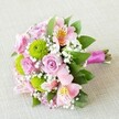 Seen from a slightly elevated angle, this radiant Bridesmaid bouquet by Flower Delivery Queensbury reflects the artistry and warmth that define local celebrations around The Broadway. Soft pink roses, at once classic and timeless, bloom at differing stages, their petals catching the soft light and suggesting delicate sweetness. Nestled among them, vibrant green button chrysanthemums stand out with their playful spherical forms, providing a lively spark that ensures the bouquet's joyful character. Pink alstroemeria, each petal traced with yellow and cinnamon-brown streaks, introduce movement and a touch of exotic refinement reminiscent of special occasions in Queensbury's grand halls and gardens. Sprigs of cloud-like white baby's breath interlace between the featured blooms, imparting a weightless, airy texture that elevates the arrangement's romantic and celebratory mood. Deep green leaves arc and curl at the edges, offering a lush, grounding frame suited to the elegance of a wedding day. The stems, precisely aligned, are cinched with a brilliant magenta satin ribbon, its sheen glowing in the light and forming a handle fit for a proud bridesmaid. The exposed natural stems hint at authenticity and freshness, while subtle fabric texture beneath adds comfort and homeliness. Exuding happiness and grace, this bouquet is perfect for sharing heartfelt moments-from wedding aisles in Queensbury to anniversary surprises-showcasing the vibrancy, local pride, and skilled hands that bring each flower to brilliant life.