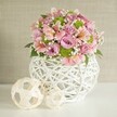 A fresh spring-inspired floral arrangement sits gracefully atop a shining white surface, glowing softly under gentle light. The bouquet features plush pastel roses in pink and lavender, their sumptuous petals layered and full, accompanied by sprightly lime green button chrysanthemums for a pop of modern vibrance. Pink alstroemeria, each with slender petals and subtle markings, weave throughout the display, bringing playful movement and quiet elegance. Sprigs of feathery white baby's breath nestle between blooms, infusing the design with a light, almost whimsical air, while scattered deep green leaves add depth and natural beauty. The vase-an eye-catching, rounded vessel of white, intricately woven strips forming an open lattice pattern-evokes both contemporary artistry and rustic charm, echoing the taste of Queensbury's stylish residents. Alongside, two decorative balls crafted from natural-toned woven material-one large, one small-introduce visual balance and subtle continuity, reminiscent of handmade pieces found in local boutiques or crafts fairs around Mollison Way. The light, textured beige wall in the background frames the scene with calming neutrality. This arrangement exudes warmth, serenity, and creative spirit, making it a perfect centerpiece for weddings, special occasions, or simply brightening a Queensbury home. Every element speaks of thoughtful local floristry and an appreciation for both beauty and craftsmanship.