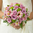 In this photograph, a romantic bridal bouquet is captured as if just finished on the workbench of a Queensbury florist, every flower placed with care and meaning. At the centre, dusky mauve and lilac-toned roses form a lush cluster, their cupped, velvety petals unfolding in tight, spiralling whorls that speak of trust, tenderness, and enduring partnership. Scattered among them, radiant pink alstroemeria stretch outward like butterfly wings, each bloom marked with soft speckles and streaks of yellow and rose, giving the arrangement a sense of openness and lively movement. Small, rounded green button chrysanthemums nestle playfully between the larger blooms, their bright chartreuse colour and textured, cushion-like heads offering a refreshing counterpoint to the gentle pastels. Fine sprays of gypsophila thread through every gap, adding a delicate veil of white, like a breath of mist drifting above the flowers. Only the most polished stems and glossy green leaves are visible, trimmed so the bouquet appears abundant yet effortlessly light in the hand. The flowers sit in soft, natural daylight, as though resting near a window overlooking one of Queensbury's quiet residential streets, their colours gently enhanced by the illumination. You can almost imagine the subtle mix of fragrances-powdery rose, green floral notes from the chrysanthemums, and a crisp leafy freshness-coming together in the air. This hand-tied bouquet feels perfectly suited for a wedding at a local venue or an intimate celebration at home, an arrangement that reflects both thoughtful craftsmanship and the gentle optimism of a new chapter shared here in Queensbury.