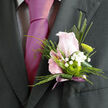 This boutonnière is captured as if it has just been carefully pinned to a groom's lapel somewhere near Queensbury Station, composed with the quiet confidence of an experienced wedding florist. At its centre sits a pale blush-pink rose, petals velvety and delicately cupped, holding a sense of anticipation and tenderness. Around the rose, slender leaves and polished greenery form a graceful frame, while tiny white gypsophila blossoms scatter like soft stars, bringing lightness and an airy, cloud-like texture. Nestled against the rose is a tight green button chrysanthemum, crisp and contemporary, its rounded form adding a modern accent that plays beautifully against the romance of the rose. The stems are wrapped with a muted mauve ribbon, tied smoothly and allowed to trail just enough to suggest movement and celebration rather than formality. The colour palette-blush, fresh green, and soft lavender tones-feels perfect for a wedding at a local Queensbury venue or a formal celebration at a nearby restaurant. Imagining the faint floral scent, you can almost hear the murmur of guests and the click of cameras as the wearer greets family along Church Lane or heads towards a registry office. Every element in this small piece speaks of care: the neat finishing, the balance between traditional rose and modern chrysanthemum, and the delicacy of the gypsophila, all coming together in a boutonnière designed to endure a full day of memories while looking effortlessly elegant.