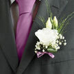 A single wedding boutonnière rests against a dark, finely cut lapel, its design speaking of quiet confidence and thoughtful craftsmanship that feels right at home in Queensbury. The focal point is a white lisianthus bloom, fully open yet soft around the edges, its layered petals unfurling like silk. Under gentle indoor lighting, the bloom appears almost luminous, a tranquil contrast to the depth of the suit fabric. Nestled behind it, two elegant lisianthus buds reach upward, their smooth, tapered forms symbolising the promise of the days ahead, much like the new journeys that begin for couples marrying near Queensbury Park or along Church Lane. Delicate clusters of baby's breath surround the feature flower, creating airy pockets of whiteness that resemble tiny puffs of cloud or early-morning mist drifting across the arrangement. Among these, slender strands of variegated foliage and fine green blades add a sense of spontaneity, breaking up the soft textures with a touch of structured wildness. At the base, a rich green leaf is looped and tucked neatly, forming a natural cradle for the stems and adding depth with its polished surface. A narrow mauve ribbon is wrapped around the stem and tied into a precise bow, its colour subtly echoing a coordinating tie or pocket square chosen with care from a local boutique. The ribbon's gentle sheen reflects a hint of lavender tone, bringing a whisper of colour without distracting from the purity of the white flowers. Imagining the piece in motion, you can almost hear the soft rustle of fabric as the groom greets family and friends, the faint floral scent mingling with the cool air of a Queensbury venue. It feels like a small, wearable promise crafted by a florist who knows how to balance tradition, grace, and heartfelt celebration.