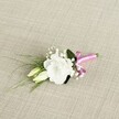 A delicately crafted wedding boutonnière lies at a gentle angle on a pale beige, linen-like fabric, its fine woven texture softly catching the light. At the heart of the design is a single, fully opened white eustoma bloom, its ruffled petals layered in smooth, velvety folds that form a graceful, rounded cup. The flower faces slightly to the right, allowing you to almost feel the softness of each petal and imagine the faint, clean fragrance often associated with fresh spring blossoms. Just to the left sit two slender eustoma buds in creamy pale green, their tips still closed, suggesting a quiet sense of promise and new beginnings that suits a wedding in Queensbury's local registry office or a celebration at a nearby country pub. Around the focal flowers, tiny white clouds of baby's breath are tucked between the stems, their miniature blooms adding airiness and a romantic, mist-like texture. Slender, dark green, grass-like blades sweep outward toward the bottom left, echoing the gentle curve of a lapel, while a single broader deep green leaf steadies the whole piece from behind, adding depth and structure. At the base, the stems are neatly gathered and bound with a vibrant fuchsia satin ribbon, tied into a jaunty little bow that introduces a joyful burst of colour against the calm neutrals. The contrast of bright pink ribbon, white petals, and rich greenery against the oatmeal-coloured backdrop evokes the feeling of a carefully chosen buttonhole waiting on a dressing table before a ceremony, perhaps moments before a groom or groomsman in Queensbury pins it to a suit jacket for a day filled with photographs, toasts, and heartfelt memories.
