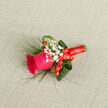 In this refined boutonniere, a vivid fuchsia rose becomes the clear focal point, its plush petals swirling in layers of deep pink that suggest both confidence and tenderness, like a small, living keepsake crafted for a Queensbury celebration. The rose is surrounded by delicate sprigs of white baby's breath, each tiny blossom forming a soft constellation of dots that seem to float around the main flower, softening its intensity. Nestled beside them, rounded scarlet hypericum berries bring a rich, berry-red contrast, their polished surfaces echoing the joyful mood of a wedding morning or prom evening. Carefully curved aspidistra leaves frame the composition, their broad, shiny greens folded into graceful loops that cradle the rose, while slender strands of ornamental grass extend outward, adding a light, almost breezy movement. The stems are tightly wrapped in a crimson ribbon, wound in smooth spirals that give a clean, tailored finish, suggesting the steady, practiced hands of a local florist accustomed to dressing grooms and guests for ceremonies across Queensbury, from church weddings off Kenton Road to civil services near the station. Set against a subtle, neutral-textured background, the rich pinks, reds, and greens seem to glow, with gentle daylight picking out each detail: the gloss of the berries, the matte softness of the petals, the faint veins in the leaves. Imagine pinning it onto a jacket, feeling the cool, fresh stems beneath the ribbon and catching a fleeting note of fresh rose and greenery, a small yet meaningful accent designed to travel through the day as a quiet emblem of joy, love, and shared memories.