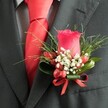 A close-up, high-angle photograph shows a meticulously crafted wedding boutonniere pinned to the left lapel of a deep charcoal suit jacket, the kind you might see on a groom stepping out for a ceremony in Queensbury Park. At the heart of the design sits a single, velvety deep red rosebud, its petals tightly furled yet richly layered, rising upright with quiet confidence. Nestled around its base are delicate clusters of tiny white gypsophila, like a soft cloud of baby's breath that gently lightens the mood and adds a romantic haze against the dark wool of the suit. Interspersed among the white blossoms are several glossy red berries, likely hypericum, their rounded shapes catching the light and mirroring the rose's crimson hue for added depth and texture. Behind this focal cluster, slender, grass-like blades of vivid green foliage fan outward, while broader, darker leaves curve protectively around the stem, giving the buttonhole structure and a natural, garden-fresh feel. A narrow red satin ribbon is neatly wrapped and tied into a small bow at the base, its sheen echoing the gleam of the rose petals and berries, and suggesting a careful, practiced florist's hand. Just beneath the lapel, a bright red tie in smooth satin or silk runs down the chest, its perfectly formed knot framed by a subtly patterned dress shirt in pale grey or white, perhaps a fine check or houndstooth weave. The combination of textures-crisp fabric, polished berries, and soft petals-creates a refined, cohesive look that suits weddings, proms, or black-tie events across Queensbury, especially when styled by a local florist who understands classic, understated elegance.
