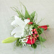 Set against a soft, pale surface that lets every colour sing, this hand-tied bouquet feels like a personal love note composed in flowers, the kind you might carry down the aisle at a Queensbury ceremony or present on a milestone evening. At its heart, a luminous white lily opens with poise, each creamy petal arching outward in a star-like shape, smooth and almost satiny to the eye. Nestled around it, rich red roses unfold with velvety depth, their tightly layered petals creating pools of shadow that hint at devotion, passion, and unspoken promises. Clusters of shiny red berries dot the spaces between blooms, bringing a cheerful pop of brightness that keeps the arrangement lively rather than solemn. Wispy sprays of gypsophila scatter tiny white blossoms throughout, like a dusting of gentle snow, softening the transition between the bold reds and the central lily. Light, airy greenery threads through the bouquet, adding subtle arcs and tendrils that give the design a sense of ease and natural movement, reminiscent of a stroll through the gardens near Queensbury Station on a calm afternoon. The stems are gathered tightly and finished with a slim band of scarlet satin ribbon, smooth and refined, signalling a special moment without overpowering the flowers' beauty. The impression is one of graceful celebration and intimate sentiment, perfectly suited for local wedding bouquets, engagement gifts, or romantic surprises delivered across Queensbury. You can almost sense the fresh, clean fragrance of lily mingling with the deeper, classic scent of rose, creating a bouquet that invites both the eye and the heart to linger.