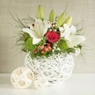 A graceful floral arrangement is displayed as if ready for a special event in Queensbury, set against a softly textured backdrop in shades of warm beige and light grey that gently fade into the distance. At the heart of the composition is a distinctive round white vase, its open-weave design formed by criss-crossing strands that resemble finely woven wicker or slender branches shaped into a contemporary basket. Overflowing from this sculptural vessel is a vibrant bouquet dominated by white lilies and red roses, accented with fresh greenery and playful textures. The lilies, some fully open and others tightly enclosed in green buds, stretch upward and outward with broad, smooth petals that curl slightly at the tips, revealing golden stamens and subtle speckling at their throats. Nestled amid their purity are deep red roses, one particularly striking bloom with a tightly coiled spiral of velvet petals forming a rich focal point. Between these timeless flowers are bright green, spherical pompons-likely green dianthus or chrysanthemums-adding a mossy, cushion-like texture that brightens the entire design. Fine clusters of tiny white baby's breath float through the bouquet, softening the lines and lending an airy, romantic quality well suited to a wedding table along Queensbury's quieter streets. Small clusters of glossy red berries peek out among the greenery, adding a gentle festive note without overpowering the arrangement. Slender fronds and darker, broader leaves spill gracefully over the rim of the vase, filling in gaps and creating a sense of lush abundance. To the left of the vase, two decorative spheres made from pale woven material rest on the smooth white surface, echoing the vase's pattern and tying the scene together with understated elegance, perfect for a refined celebration or intimate dinner party.