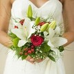 Soft morning light filters onto a bride in Queensbury, her arms cradling a bouquet that feels as though it was freshly gathered from the lush borders of Roe Green Park. Creamy white lilies blossom boldly, their petals unfurling beside rich red roses whose tightly coiled petals speak quietly of love and commitment. Delicate sprays of baby's breath drift between the larger flowers, evoking the gentle mist of a new day, while vivid hypericum berries and vibrant green orbs add playful texture and flashes of colour. The bouquet's green foliage-feathery ferns and glossy leaves-forms a lush base, inviting memories of local gardens waking to spring. Every detail feels chosen with careful intent by a skilled Queensbury florist, composing a bouquet both refined and organic. The bride's gentle grip and the subtle pattern of her gown complete a sense of serene anticipation, marking a beginning rooted in natural grace and the joyful spirit of weddings around Queensbury.