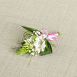A lovingly arranged white floral buttonhole is laid out against a pale oatmeal linen backdrop, the subtle weave of the fabric adding warmth and texture behind the blooms. At the heart of the composition, a fully opened white alstroemeria unfurls its petals like fine satin, each inner petal traced with delicate reddish-brown streaks that give the flower personality and gentle drama. Around it, several smaller white flowers sit in various stages of opening-some still curled as buds, others just beginning to reveal their petals-suggesting promise and anticipation, like a special day about to begin in Queensbury. Wisps of baby's breath drift among them, tiny white dots on fragile stems that soften the boutonniere's outline and create an airy, romantic haze. On the left, a distinctive, elongated cone of bright, citrus-green buds, likely ornithogalum, adds a contemporary touch and a crisp, sculptural line, balancing the softness of the other blooms. Dark green leaves spread out behind the flowers, creating a rich, glossy backdrop, while slender blades of foliage arc gently over and beyond the design, echoing the natural curves you might see in the landscaped corners of nearby parks. At the base, a polished pink satin ribbon gathers the stems into a neat, compact bundle, tied into a tidy little bow that introduces a lighthearted, celebratory accent. The piece feels at once elegant and heartfelt, ideal as a wedding boutonniere, prom accessory, or corsage for a special event in and around Queensbury, where a small spray of fresh flowers can express love, pride, or quiet joy. You can almost imagine the cool touch of the leaves and the faint, green-sweet scent rising from this handcrafted floral detail.
