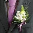 A close-up, high-angle photograph captures the upper torso of a man dressed for a formal occasion in Queensbury, his attire framed in soft, natural light. He wears a finely tailored charcoal grey suit jacket with a crisp, pale dress shirt beneath, patterned with a subtle grid that adds quiet texture. Down the centre of his chest falls a smooth, solid lavender tie, neatly knotted and lying perfectly flat, its cool sheen echoing the elegance of a spring wedding or school prom near Queensbury Park. Pinned precisely to the left lapel, the boutonniere becomes the true focal point: a tall, pale green spire of tightly closed buds rises at the top, flanked by several pristine white alstroemeria blooms, each petal brushed with fine streaks of maroon and soft gold at the throat. Clusters of airy white gypsophila, or baby's breath, weave between the flowers, creating a light, cloud-like halo. Around them, slender, grass-like blades and broader, glossy deep-green leaves arc and curve, giving the arrangement movement and a sense of freshness, as if just gathered from a local Queensbury garden. At the base, a neatly tied satin ribbon in matching lavender cinches the stems into a tidy bundle, its gentle shine mirroring the tie and bringing the colour story together. The overall impression is one of quiet sophistication and hopeful anticipation, the kind of carefully considered buttonhole a groom or groomsman might choose from a trusted local florist for a ceremony in or around Kingsbury Road, marking a once-in-a-lifetime moment with simple, refined beauty.