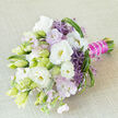 A sophisticated hand-tied bouquet is captured resting at a graceful angle on a softly textured light beige fabric, the neutral backdrop allowing every flower and ribbon detail to breathe. Dominating the composition are lush white lisianthus blooms, their silky, ruffled petals opening gently to reveal warm, creamy yellow centres, each flower at a slightly different stage of unfolding. Among them, narrow buds in pale green and ivory stand tall, promising future blossoms and giving the design a sense of quiet movement. Threaded delicately between the lisianthus, pastel lavender and blush-pink phlox form airy, cloud-like clusters, softening the structure and adding a romantic haze of colour. Striking contrasts arrive in the form of deep purple allium florets, each one a tiny star-shaped cluster forming a rounded, spiky sphere that lends contemporary flair and visual intrigue. Fresh greenery in various shades of green, from slender leaves to more architectural, spiky elements, brings depth and texture, echoing the leafy verges and gardens tucked around Queensbury's residential streets. The stems are meticulously gathered and bound in a bold magenta-fuchsia satin ribbon, wrapped securely along the handle and ornamented with a strand of petite white pearls that trace a refined, shimmering line. This detail suggests the careful hand of an experienced florist, the sort you might find at Flower Delivery Queensbury preparing arrangements for weddings at local venues or intimate celebrations at home. The bouquet's palette of whites, soft purples, and greens radiates freshness and tenderness, evoking a calm morning sky and the scent of newly opened flowers. It feels ideal for a bride stepping out of a car on a quiet Queensbury lane, a milestone anniversary, or any special occasion where elegance, love, and personal attention to detail are at the heart of the moment.