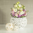 A rounded, lush bouquet sits like a quiet sunrise on a smooth white shelf-a graceful mix of cream and soft-yellow Eustoma (lisianthus) whose layered petals open like whispers. Around them, soft pink Stock forms gentle halos of fragrance and texture, while compact, starry violet Allium punctuate the arrangement with a crisp, sculptural edge. A ring of darker green leaves hugs the base of the stems, neatly gathering the blooms and giving the silhouette a polished, corseted look. The flowers are housed in a distinctive spherical vase-an airy white orb braided from slim branch-like twigs-its open latticework allowing glimpses of green stems and suggesting craftsmanship as much as function. Two handwoven rattan-like orbs rest nearby on the same bright surface; their pale wood tones mirror the vase's natural weave and bring a subtle warmth against the cool pastels. Soft, diffused light from a nearby window bathes the scene, throwing gentle reflections across the table and emphasizing the linen-like texture of a neutral greige wall behind. The scent would be delicate-green, slightly sweet, a whisper of musk-matching the bouquet's understated mood. Imagine this arrangement brightening a Queensbury kitchen table or calming a bedside in a home not far from Kingsbury and Wembley: it's an offering of quiet elegance, crafted to soothe, uplift, and fit seamlessly into a modern neighbourhood interior.