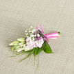 Resting on a subtly woven, neutral cloth, this refined boutonnière appears as though it has been paused in a moment of quiet anticipation, ready to be pinned to a jacket for a Queensbury celebration. Creamy buds of freesia, their petals crisp and slightly translucent, cluster at the centre, promising a soft, sweet fragrance that suggests fresh mornings along Church Lane. Around them, pale blush-pink flowers open with a tender, sigh-like softness, each petal curved and smooth, creating a romantic interplay of ivory and pastel. Tiny tufts of gypsophila, or baby's breath, rise delicately above the main blooms, like miniature puffs of cloud adding texture and airiness without overwhelming the composition. Slender blades of greenery arc gently outward, while broader green leaves curl inwards, giving the design both structure and movement, a balance between wild garden charm and considered florist artistry. The stems are wrapped in a satiny pink ribbon, its sheen catching the soft, natural light, and the knot is tied with precision, suggesting experienced hands at work, perhaps in a busy local studio near Queensbury station. The palette is calming-ivory, blush, and fresh green-ideal for a wedding lapel, a civil ceremony at a nearby registry office, or a milestone celebration at a family gathering. Though small, the boutonnière feels complete and intentional, a personal floral accent that adds a brush of colour and an intimate connection to nature. Its overall impression is one of gentle romance, understated sophistication, and the kind of thoughtful detail that guests will quietly admire throughout the day.