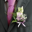 A close-up photograph focuses on the left lapel of a sharply tailored charcoal suit, the kind you might see on a groom arriving at a Queensbury wedding venue near the village green. The fabric of the jacket shows a fine, almost brushed texture, with the stitching on the lapel crisp and precise. Beneath the jacket, a pale dress shirt with a subtle grid pattern peeks through, adding a soft contrast to the rich, solid magenta tie that glows with a silky sheen in the gentle light. Pinned neatly to the lapel is a carefully crafted buttonhole, its flowers arranged with the attentive eye of a local florist who knows how to balance colour and form for a special day. At the top, slender cream and white buds, reminiscent of snapdragons, stand upright and unopened, suggesting a moment of anticipation. Just below, pastel pink and light lavender blooms cluster together in a rounded, romantic posey, suggesting sweet peas or delicate stock, their petals appearing soft enough to crush between your fingertips. Tiny clouds of white gypsophila weave through the arrangement like a light mist, punctuated by a few deeper purple florets that add depth and elegance. Broad, glossy dark green leaves cradle the flowers, while feathery sprays of brighter green foliage arc outward, giving the buttonhole movement and a touch of wild garden charm, similar to the greenery you might see along the paths around Queensbury Park. At the base, a slender lavender satin ribbon is tied in a precise bow, its gentle shine echoing the tie and uniting the colours. The overall mood is one of quiet confidence, joy, and thoughtful coordination-a single, elegant detail ready to be worn at a wedding ceremony or formal celebration in and around Queensbury.