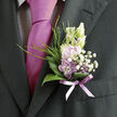 This boutonnière rests gracefully against a dark, well-tailored suit, a small but striking focal point that could easily belong to a groom stepping out of a car on a Queensbury side street or a guest arriving at a celebration near the local green. Soft lilac petals form the main bloom, their pastel tones gently transitioning from a dusky lavender at the base to a slightly lighter, almost powdered hue at the tips. Each petal looks thin and tender, like fine tissue paper, yet purposeful in its shape. Surrounding this central flower are clusters of tiny white gypsophila, creating an airy halo of texture that feels like morning mist hovering over a meadow. Shiny, curved garden leaves cradle the composition, deep green and almost waxy in appearance, their smooth arcs framing the lilac and white. Threading through these leaves, a few slender blades of vivid green grass stretch upward, adding height, movement, and a hint of natural spontaneity-as though the flowers were freshly gathered from a countryside path not far from Queensbury's quieter corners. The stems are bound carefully with a narrow lavender ribbon, tied snugly at the base with a soft sheen that reflects gentle indoor light and pairs beautifully with the nearby tie. The result is an arrangement that speaks of understated elegance and thoughtful design, with a fragrance you might imagine as light, sweet, and clean. Crafted in the intimate scale of a single lapel, this boutonnière showcases the skill of a seasoned florist who understands balance, romance, and the importance of detail for weddings and formal events across Queensbury.