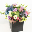 A striking floral display bursts with colour and texture from within a modern, square matte black box, thoughtfully composed by a florist rooted in Queensbury's vibrant community. The arrangement showcases luxuriant clusters of deep indigo and soot-blue hydrangeas, their full, cloud-like heads catching the daylight and forming a textural heart to the bouquet. Intertwined amongst them are soft lavender roses and blush-pink roses, their petals unfolding with delicate grace, echoing the romance of local garden walks along Kingsbury Road. Accents of creamy white lisianthus, with their ruffled, trumpet-like blooms and closed buds, add a refined brightness, while sprigs of lively green-possibly 'Green Trick' dianthus-lend a fresh, playful counterpoint. Wispy pink astilbe sprays introduce a whimsical softness, hovering above glossy foliage and scattered new buds that peek through, suggesting a freshly gathered garden arrangement. Set against a gleaming white backdrop and illuminated by even, natural lighting, the bouquet's colours seem to glow-purples, blues, pinks, and creamy greens all harmonising in serene celebration. The sleek black container imparts a touch of urban sophistication, perfect for modern homes and stylish offices across Queensbury. Designed for occasions of affection, appreciation, or congratulations, this bouquet is as versatile as it is beautiful, offering a radiant presence and a sense of local pride to every recipient.