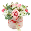 A romantic hatbox arrangement is showcased against a crisp white backdrop, every detail carefully styled to draw attention to the flowers. The round gift box is a soft pastel pink with a smooth, matte surface that feels calm and contemporary, much like the elegant homes scattered around Queensbury. Around its middle, a slender coral-red satin ribbon is tied into a neat bow, adding a bold touch of colour and movement. A small cream gift tag with subtle green writing hangs from the bow, quietly suggesting a personal message or dedication. From the open top of the box, a lush, rounded bouquet spills out, creating a full, domed silhouette. At the centre, luxurious pink roses unfurl their velvety petals, some in pale blush, others tipped with deeper pink, capturing that moment of perfect bloom. Surrounding them, clusters of crisp white daisy-like chrysanthemums with fresh green centres bring brightness and texture, balancing softness with a hint of structure. Scattered amongst these are smaller intense pink blossoms-likely spray roses or miniature carnations-that add little sparks of colour, like joyful notes in a melody. Sprigs of eucalyptus in silvery green curve gently around the blooms, their rounded leaves contrasting with the petal shapes, while tiny brownish seed pods peek through, giving an organic, slightly wild finish. The overall look is polished yet inviting, a bouquet that feels at home as a birthday surprise, an anniversary gift, or a simple thinking of you gesture. It is easy to imagine this pastel hatbox arriving by local flower delivery to a doorstep in Queensbury, bringing fragrance, softness, and a sense of occasion into someone's day.