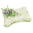 A tender, rectangular sympathy pillow artfully composed from hundreds of small, pristine white chrysanthemum heads, photographed on a clean white backdrop with a soft shadow that hints at its gentle, puffy profile. The chrysanthemums are densely clustered to form a uniformly plush, almost cloud-like surface that speaks of quiet rest and purity; you can almost sense the soft, papery texture and the faint, green-tinged scent typical of freshly cut chrysanthemum. Around the entire perimeter a light lime-green ribbon is carefully pleated into neat loops, its satiny sheen providing a subtle, fresh contrast that defines the pillow's softened corners. Anchoring the piece across the upper-left corner is a composed cluster of accent blooms: layered white lisianthus with their delicate ruffles, deeper purple lisianthus in both open florets and tight buds, and slender swathes of feathery fern and glossy green foliage that add depth and an honest, botanical freshness. A darker green leaf wrap holds the stems together at the base of the spray, visible as an elegant, natural binding. The composition is lit with soft, even light that emphasizes texture and shadow without distraction, suggesting the care of a neighbourhood workshop near Queensbury Station and the peaceful green of nearby Fryent Country Park. Intended as a respectful graveside or memorial tribute, this arrangement conveys solace, dignity and the quiet care of a local florist: a floral embrace for those remembering a loved one in Queensbury, Kingsbury, or nearby Wembley, crafted to comfort and give shape to sorrow when words feel inadequate.