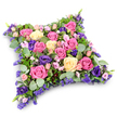 A graceful, pillow-shaped floral tribute is presented against a clean white backdrop, appearing as a gently rounded cushion completely covered in fresh blooms. The colour palette is soft yet rich, blending warm pinks and creamy yellows with cool purples and abundant greenery to create a comforting harmony. At the heart of the design, full-bloom roses in shades of deep fuchsia, pastel pink, and pale buttery cream unfold their layers of petals, each flower perfectly opened to show its intricate centre. Interspersed between them are velvety purple lisianthus, whose multi-petalled, trumpet-like blooms introduce depth and a slightly more formal note, reminiscent of traditional memorial flowers. Smaller spray roses, delicate and romantic in light pink, nestle close to clusters of tiny lavender-toned filler flowers, likely statice or limonium, which lend a fine, lacy texture and fill any remaining space so the cushion appears plush and complete. Beneath and around the blossoms, a mixture of foliage adds structure: round, silvery eucalyptus leaves bring a cool, soothing green, while darker, slender, fern-like fronds add contrast and subtle movement. The cushion's softly curved corners and gently domed surface evoke the idea of rest and peace, a floral pillow on which memories can quietly settle. Set against the stark white background, the colours stand out with particular clarity, as though the tribute has just been placed on a stand at a chapel of rest or family home in Queensbury, not far from the calm streets leading toward Canons Park. This arrangement feels both tender and composed, a heartfelt way to express sympathy, gratitude, and lasting love at a funeral, memorial, or graveside farewell.
