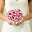 This bridal bouquet rests gently in the bride's hands, a perfectly rounded cluster of blush-pink roses framed by a soft veil of white gypsophila. Each rose is full and velvety, with petals spiralling out from the centre in subtle gradients of blush and ivory, as if painted with the faintest watercolour wash. Sprays of baby's breath weave between the roses and around the outer curve, their tiny flowers forming a cloud of fine texture that lightens the bouquet's silhouette and gives it a dreamy, romantic haze. The entire arrangement sits snugly against an intricate ivory lace wedding dress, the gown's delicate pattern mirroring the tiny blossoms of the gypsophila. Natural, diffused daylight-similar to the gentle light spilling over gardens in Queensbury-bathes the scene, enhancing the soft glow of the pink roses and casting faint, tender shadows across the bride's hands. There is an air of serene anticipation, as if the bouquet is being held just moments before walking down the aisle in a local church or registry office near Queensbury station. One can almost sense the fragrance: fresh rose petals, lightly sweet and slightly dewy, blending with a hint of crisp greenery. The design feels at once classic and deeply romantic, thoughtfully arranged so every bloom appears cherished. Created by florists who understand both tradition and modern bridal style, this bouquet becomes more than a floral accessory; it is a quiet companion to the vows, carrying with it love, promise, and the soft joy of a Queensbury wedding morning.