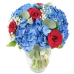 A lush, spherical bouquet sits in a clear glass vase, its voluminous sky-blue hydrangeas forming a soft, cloud-like foundation that seems to glow against a clean white backdrop. The hydrangea clusters are dense and saturated, each tiny floret overlapping the next to create a plush, tactile surface that reads cool and refreshing to the touch. Punctuating that blue expanse are deep, velvety red roses-rich in tone and with tightly furled petals that offer a passionate, classic counterpoint to the hydrangeas' airy presence. Sprigs of delicate white baby's breath peek through like fleeting stars, lending an ethereal lightness that keeps the composition from feeling heavy, while rounded, muted-green eucalyptus leaves thread through the stems and blooms, adding a soft herbal scent and an elegant, organic silhouette. The transparent vase, slightly rounded at the base and narrowing toward the top, reveals green stems submerged in clear water, a visible sign of freshness and careful arranging. Bright, even lighting emphasizes the saturated blues and reds and the tiny textures of each petal, creating a crisp, contemporary image that still feels warm and intimate. As a local florist describing this piece from Queensbury, I picture it sitting on a sunlit table near Queensbury Station or on Honeypot Lane, brightening a kitchen or dining room with a confident, celebratory air. Whether given for an anniversary, a birthday, or simply to mark a small domestic triumph, the bouquet's combination of cobalt hydrangeas, scarlet roses, soft baby's breath and smoky eucalyptus reads as both joyful and timeless, perfectly suited to quiet moments and happy gatherings alike.