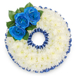 A bespoke funeral tribute crafted in the distinct shape of the number '0', photographed on a crisp white background to highlight its precise form and texture. The foundation is a dense, continuous carpet of small white spray chrysanthemums - hundreds of tightly clustered florets whose tiny petals create a soft, pillowy surface that reads as purity and calm. Around both the outer edge and the inner void a deep royal blue trim runs like a ribboned frame; the border is finely pleated satin or dyed flora, ruffled to form a subtle scallop that reinforces the wreath's circular geometry and lends a formal, ceremonial finish. On the upper left curve a trio of royal blue roses commands attention: their petals unfurl from tight centres in a rich, saturated blue that suggests serenity and reverence. Glossy dark green leaves sit just behind those roses, their sheen and vein patterns giving depth and a natural contrast to the immaculate white and vivid blue. The overall feeling is one of respectful restraint - cool, elegant, and carefully balanced - intended as a personalised numerical tribute for a memorial service, wake, or graveside farewell. As a local florist familiar with Queensbury's chapel services and community traditions, I picture this piece arriving at a small chapel or family home in Queensbury, to be placed as part of a numbered floral tribute marking an age or milestone. The satin's sheen, the chrysanthemums' soft texture, and the bold blueness of the roses together speak of honour, quiet strength, and enduring remembrance.