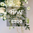 A stunning, lush floral spray in pristine white and cream tones sits against a soft, light background, its elegant pose suggesting a bridal or ceremonial centerpiece. The arrangement is built around a core of soft white roses, some still in bud and others open to reveal velvety petals, with voluminous clusters of creamy hydrangeas adding cloud-like fullness. Delicate white sweet peas or similar cascading blossoms trail downwards, their slender stems lending movement and a sense of quiet grace. Sprinkled throughout are Nigella Love-in-a-Mist, with their intricate green centers and feathery seed heads, punctuating the display with architectural texture. The whole composition rests among a generous selection of greens: broad, dark leaves form a rich backdrop, lighter variegated foliage offers gentle contrast, and fine, feathery fronds soften the edges. Rustic woody stems peek from the base, giving a natural, trailing spray that completes the horticultural sculpture. A crisp white rectangular frame overlays the central bouquet, carrying the text Florist Choice in a flowing script, CHOICE SPRAY in clear uppercase, and at the bottom, a bold price of £200. This image exudes high craftsmanship, a premium white spray suited for remembrance, celebration, or sophisticated home decor. In Queensbury and beyond, Flower Delivery Queensbury offers this as a same-day or next-day option for customers seeking a refined, gift-ready bouquet, with the promise of fresh stems and careful handling. The overall effect is serene, elegant, and versatile, a testament to timeless floristry that transcends seasons and occasions.