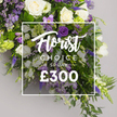 A long, elegant funeral spray or memorial tribute in a sophisticated palette of purples, whites, and greens. The lush design presents clusters of pristine white roses and soft white lisianthus, contrasted with deep purple blooms that include tall spires of delphinium or larkspur and smaller lavender blossoms such as freesias. Abundant green foliage, from ferns to eucalyptus like leaves, provides a rich backdrop and adds depth and volume. The arrangement is styled to lie flat on a casket or serve as a substantial tribute, its elongated shape enhanced by a soft neutral grey background that makes the colors pop. Overlaid on the image, slightly off center, is a semi transparent white rectangle containing text. The upper line reads Florist Choice Special in an elegant script, positioned directly above the price ?300 in bold sans serif. The layout communicates a premium bespoke option from a local florist and is suitable for sympathetic ceremonies, funeral tributes, or a dignified remembrance.