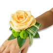 An intimate wrist corsage rests gently on the inner wrist, its centerpiece a single, fully opened rose in a soft peach to light apricot tone that reads like a warm sunrise on the skin. The rose's petals spiral inward with the fine, velvety texture you want to touch; they catch the light so the innermost folds glow while outer petals show delicate shadowing. Nestled just below and to the left of the rose are two lively lime-green button chrysanthemums - tight, spherical pom-poms whose clustered florets create a cheerful, almost tactile contrast to the rose's smooth layers. Around these blooms a base of deep, glossy green foliage - likely salal or ruscus - fans out in broad leaves to give the corsage weight and a fresh, living frame. A variegated leaf, folded and ribboned in a deliberate arc with pale striping against darker green, sweeps from beneath the rose and adds an architectural, contemporary line that guides the eye along the wrist. The floral pieces are wired and tucked so the fastening is hidden, letting blossoms appear to float on the skin.
As a florist who dresses wrists for weddings, proms and community events around Queensbury and nearby Wembley, I arrange colour and texture so a small piece can feel important: scented with the faintest rose perfume and the green, grassy whisper of leaves, this wristlet reads as romantic yet modern. Worn in soft daylight before a ceremony or beneath evening hall lights, it signals celebration - a delicate botanical hug for the person wearing it that complements formal attire and local traditions alike.