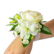 A delicately composed white wrist corsage rests gently on a fair-skinned wrist, captured against a clean white backdrop in soft, diffused light. At its heart sits a creamy ivory rose, petals unfolding with a velvety, almost luminous softness that catches the light and suggests quiet refinement. Just below and slightly left of the rose, a larger trumpet-shaped white bloom-reminiscent of alstroemeria with a subtle green-yellow throat-adds sculptural depth and a whisper of spring lime at its centre. Scattered around these main blooms are tight green buds, hinting at future opening, and airy sprays of tiny white Gypsophila that create a cloudlike halo, lending the piece an ethereal, romantic texture. Near the wrist, a cluster of small daisy-like white florets with bright golden centres provides a sunny, delicate counterpoint to the creamy tones. Deep, glossy green leaves with a smooth, waxy sheen frame the composition, their rich colour giving the whites a crisp, living contrast. The arrangement is secured by a chic, multi-strand pearl bracelet-elastic and strung with uniformly round, lustrous pearls whose cool smoothness rests against the skin and complements the blooms. The overall mood is one of timeless elegance and tender celebration, perfect for weddings, proms, or formal galas. As a Queensbury florist might note, the quiet freshness of the flowers calls to mind morning walks near Queensbury station and local green spaces; the corsage feels like a wearable promise of joy, crafted with careful hands and a thoughtful eye for texture, scent and proportion.