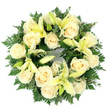 Seen from above on a clean white background, this circular sympathy wreath reads as a quiet, living circle of comfort. The design centres on a densely packed ring of creamy ivory roses - roughly fifteen to twenty blooms - whose tightly swirled petals form soft, luminous cupped faces. Interwoven with the roses are pale lemon-yellow lilies; some open wide in graceful trumpet shapes revealing pale stamens, others linger in delicate bud and half-open stages, adding gentle rhythm and the promise of unfolding life. The floral cluster sits upon a deep, verdant bed of foliage: feathery fern fronds offer a soft, lace-like edge while broader sprigs of rounded leaves - likely eucalyptus or a similar silvery-green leaf - give weight and contrast, their cooler tones pushing the creams and yellows forward. Light falls evenly from above, catching the creamy petals and creating a subtle, tender glow against the stark white backdrop. The arrangement is meticulously even, its perfect circle suggesting continuity and remembrance; it feels both serene and dignified, a classical tribute suited for funerals, graveside services, or quiet memorial moments. If you close your eyes you might imagine the roses' powdery sweetness balanced by a faint green, citrus hint from the lilies, and the cool, leafy scent of eucalyptus threading through the air. Crafted with thoughtful restraint, the wreath's palette of white, pale yellow and greens communicates peace and gentle condolence, a respectful visual embrace to honour a cherished life in Queensbury and the surrounding neighbourhoods.
