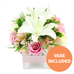 A sophisticated lily and rose arrangement is carefully styled in a square glass vase and set against a clean white background, giving it a serene, gallery-like feel. At the centre, two large Oriental lilies open generously, their white petals curling back with a soft sheen, revealing upright, dark reddish-brown stamens that add contrast and a touch of drama. Framing these focal blooms, a ring of pink roses in varying stages of blossom-from small, promising buds to fully unfurled, multi-petalled flowers-creates a sense of depth and gentle movement. Threaded through the roses are delicate sprays of alstroemeria in shades of pink and white, some petals brushed with fine yellow markings, which lend a slightly playful, garden-picked accent. Wisps of airy white baby's breath are tucked between the larger flowers, giving an impression of lightness, as though the bouquet breathes. A mix of vibrant green foliage surrounds everything, from broad, shiny leaves that catch the light to finer, fern-like greens that soften the edges, much like the mix of manicured and natural planting seen around Queensbury's residential greens and nearby parks. The flowers are anchored in a clear, square glass vase, its transparent sides revealing neatly arranged stems, while a pale pink satin ribbon encircles the centre of the vase in a smooth horizontal band, adding a refined, gift-ready touch. In the bottom right corner, an orange circular graphic with white block text clearly reads VASE INCLUDED, signalling that the glass container arrives with the bouquet. The overall mood is one of fresh elegance and quiet celebration, ideal for same-day delivery from Flower Delivery Queensbury to mark birthdays, anniversaries, or tender thank-you moments throughout the local area.